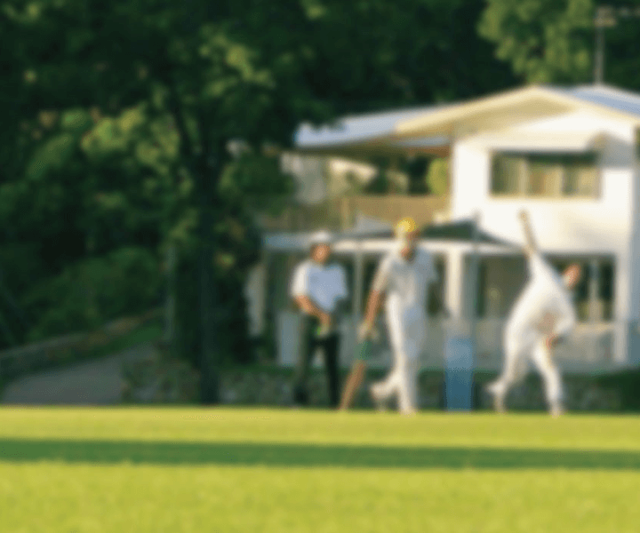 Blurred full colour image of three men playing cricket in the foreground with a white club house and trees in the background